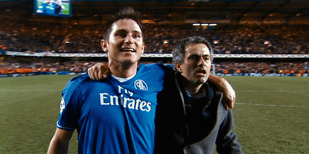 Two men celebrate with arms around each other on a soccer field, wearing team and coaching attire, with a cheering stadium crowd in the background during a night match.