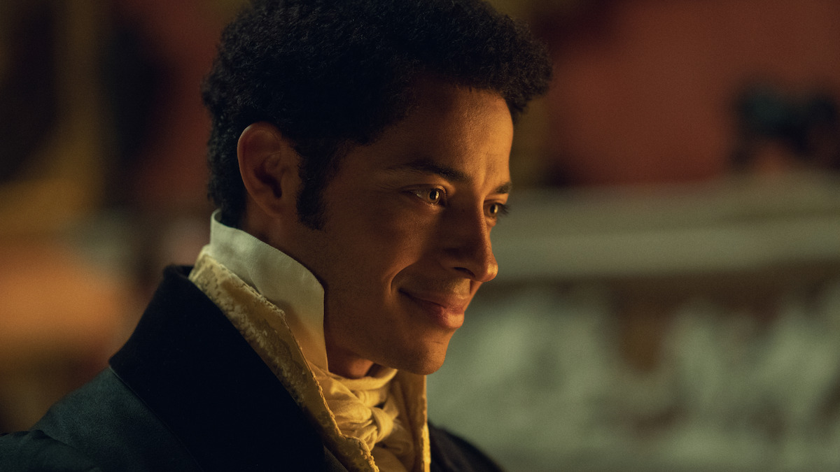 A man dressed in historical formal attire smiles warmly in a softly lit, elegant interior, likely from a period drama setting.