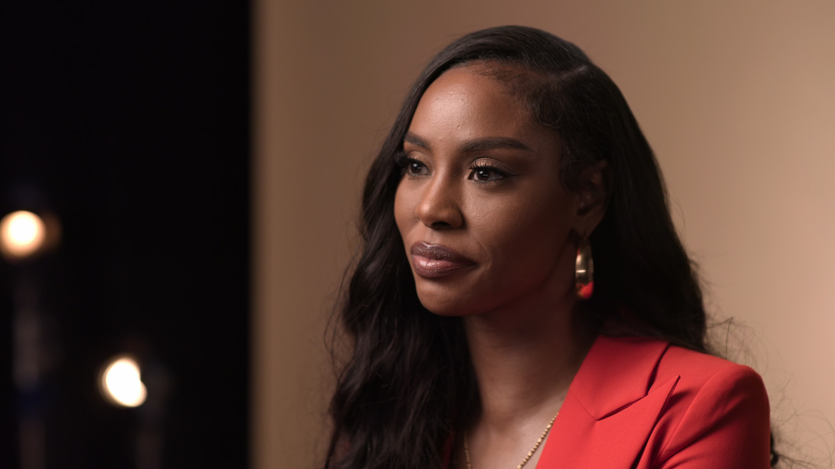 Woman with long dark hair in a red blazer sits in a softly lit studio, with a neutral background and out-of-focus lights in the distance. She appears thoughtful and composed.
