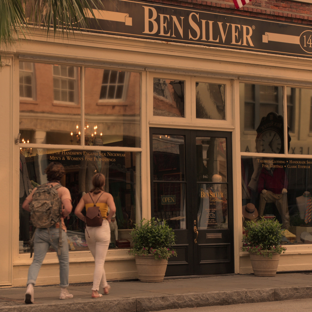 Duas pessoas andam perto da fachada da loja da Ben Silver, que tem grandes janelas com reflexos, plantas em vasos e manequins vestidos dentro da loja.