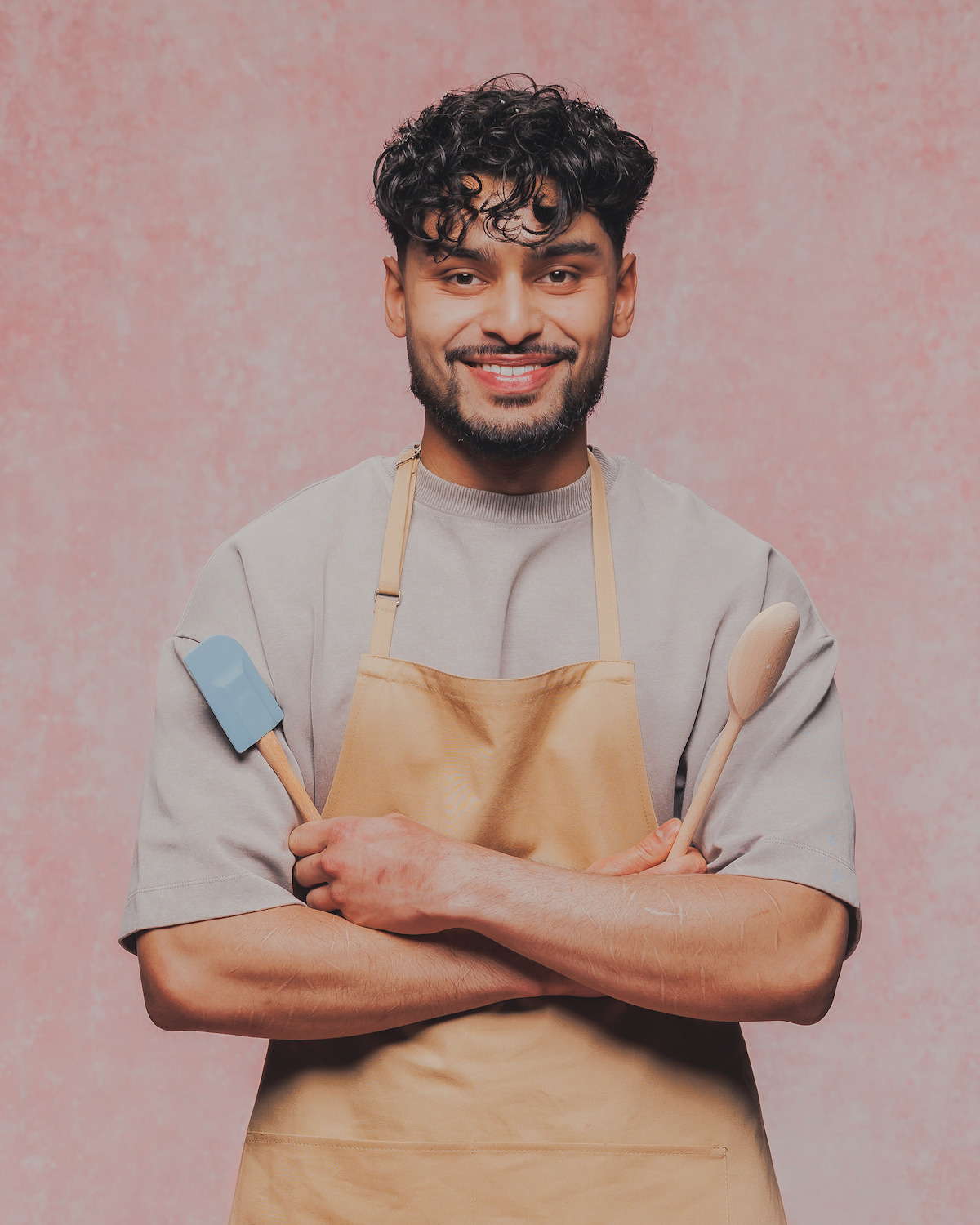 Man wearing a tan apron stands in front of a pink textured background, smiling and holding a spatula and wooden spoon, suggesting a cooking or baking setting.