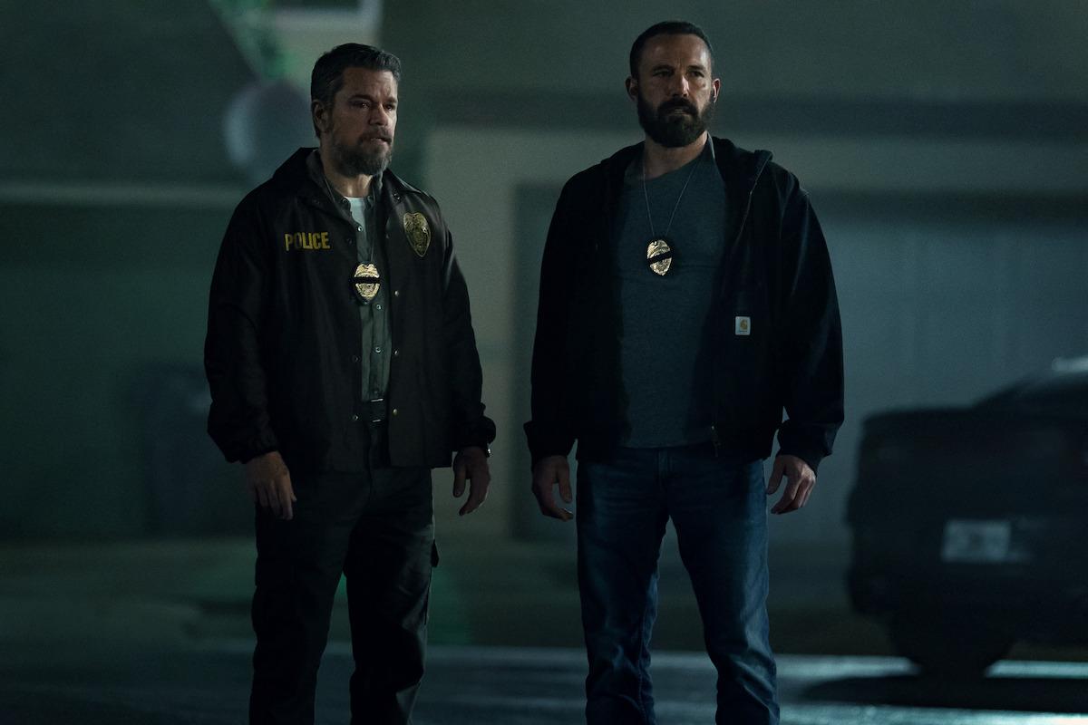 Two male police officers with badges stand in a dimly lit outdoor urban setting at night, near a car and a building, appearing serious and alert.