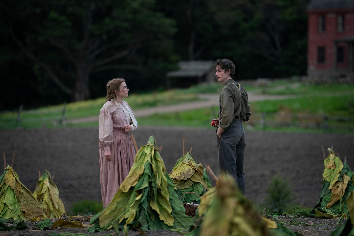 A woman and man stand in a rural field among bundles of large green leaves, appearing to talk. The scene is set outdoors with a distant red building and trees in the background, evoking a historical or agricultural atmosphere.