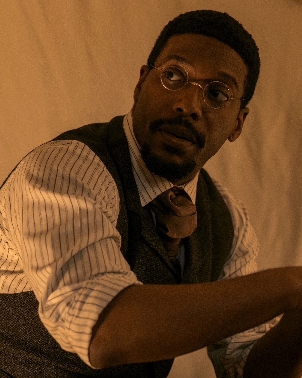 A man wearing round glasses, a striped shirt, vest, and tie sits indoors in warm, soft lighting, appearing thoughtful or in conversation, against a plain background.