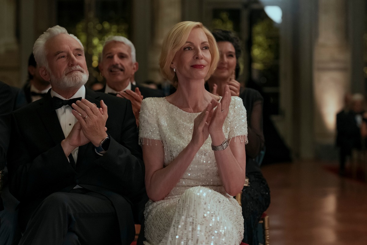Elegant people in formal attire sit and applaud at an indoor event, possibly a gala or ceremony, with warm lighting and ornate decor in the background.