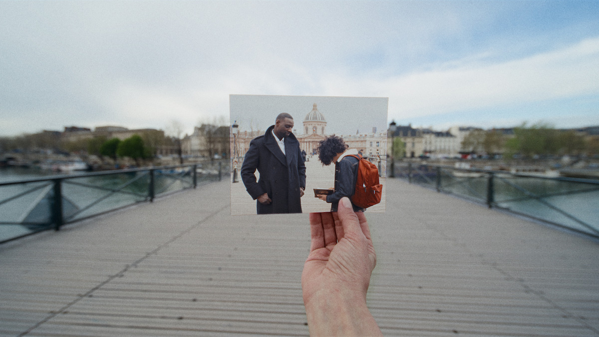 A hand holding a photo of two people on a bridge, one with a backpack, matching the real bridge in the background with buildings, river, and cloudy sky.