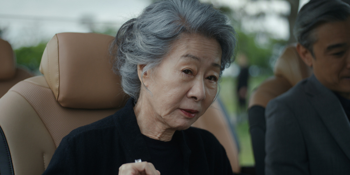 Elderly woman with gray hair sitting in a car, wearing a black top, looking slightly toward the camera, with a man beside her. The car interior is tan, and greenery is visible outside the window.