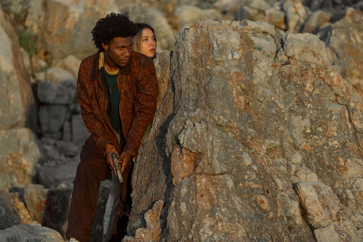 Two people hiding behind large rocky boulders outdoors; one person is crouched holding an object, both looking alert as if watching or waiting for something in a rugged, natural environment.