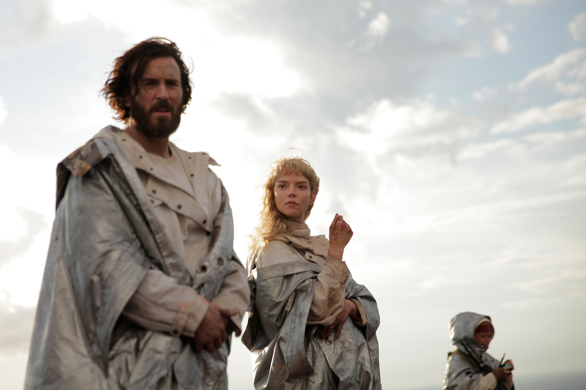 Two people in metallic cloaks stand outdoors under a cloudy sky, with another person in the background, suggesting a futuristic or sci-fi setting.