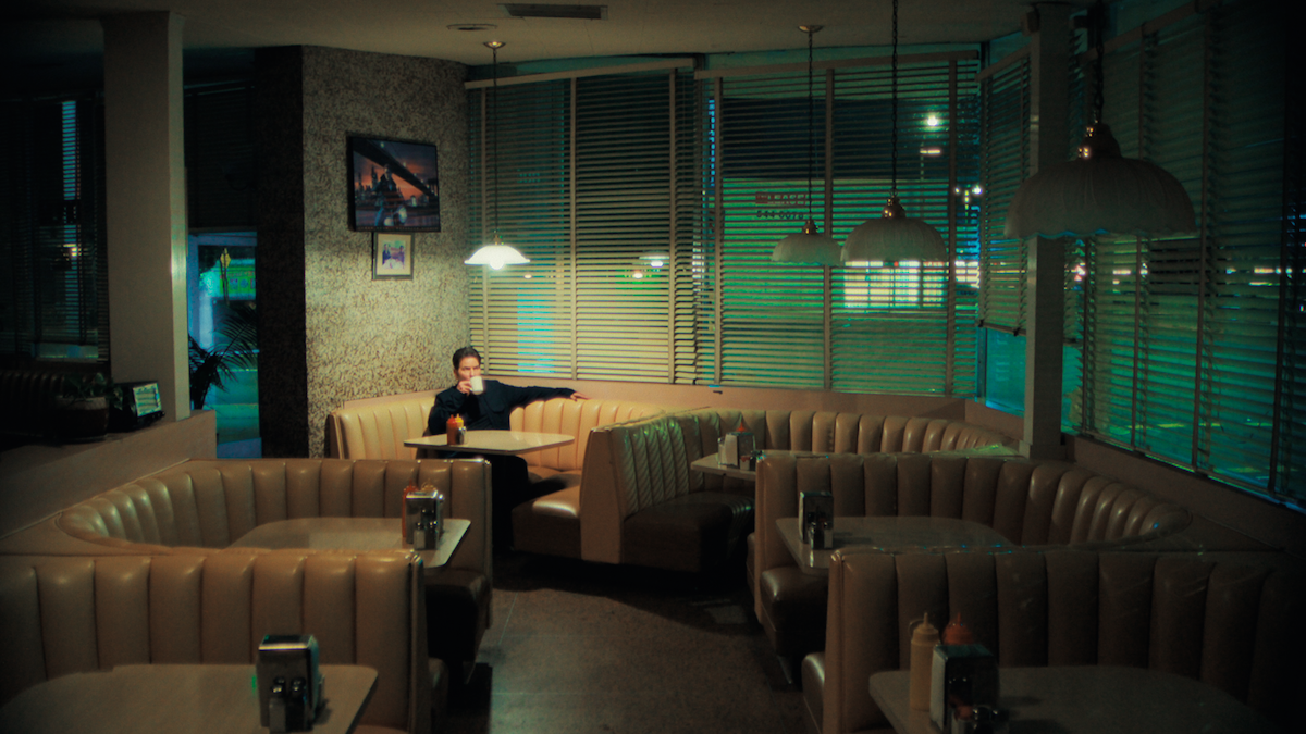 A person sits alone in a dimly lit, empty diner booth at night, surrounded by beige booths, tables with condiments, and large windows showing city lights outside.