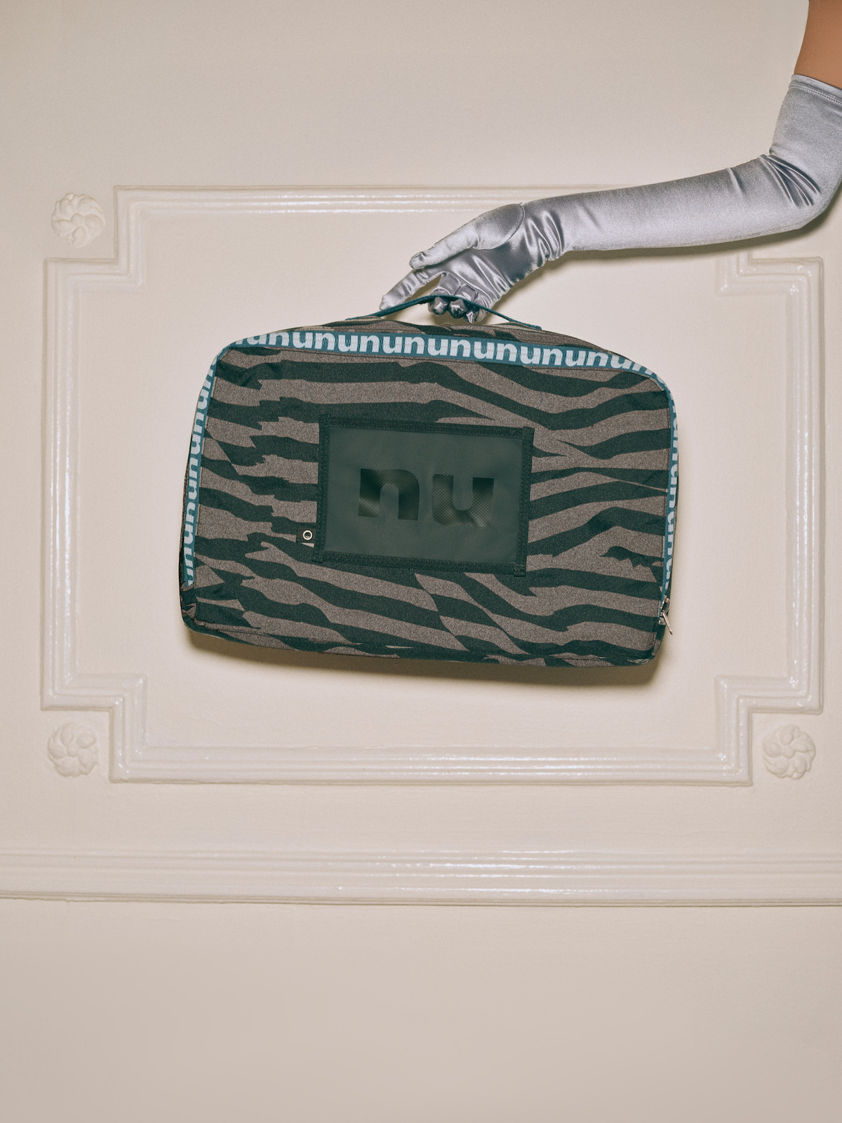 A person wearing a silver glove holds a rectangular zebra-patterned bag against a cream-colored ornate wall, highlighting the stylish accessory in an elegant indoor setting.