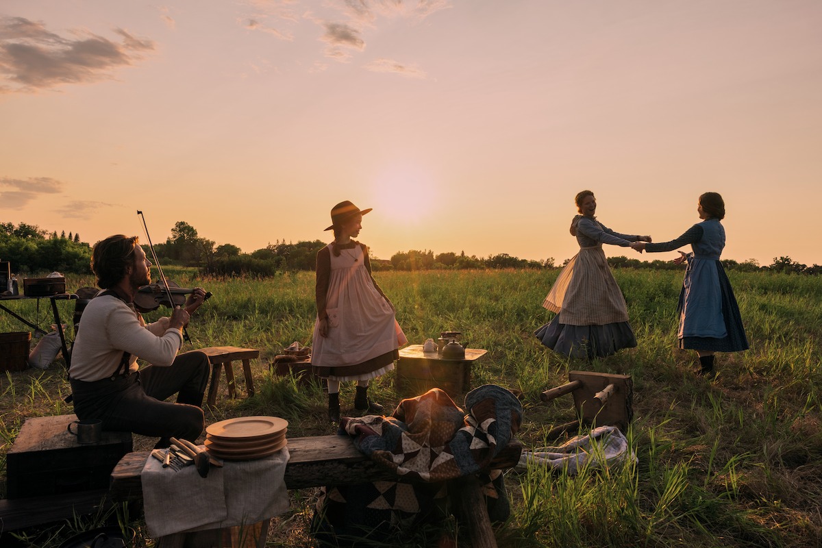 People in period clothing enjoy a picnic at sunset in a grassy field; a man plays violin while two women dance and another stands nearby, with rustic wooden benches and dishes in the scene.