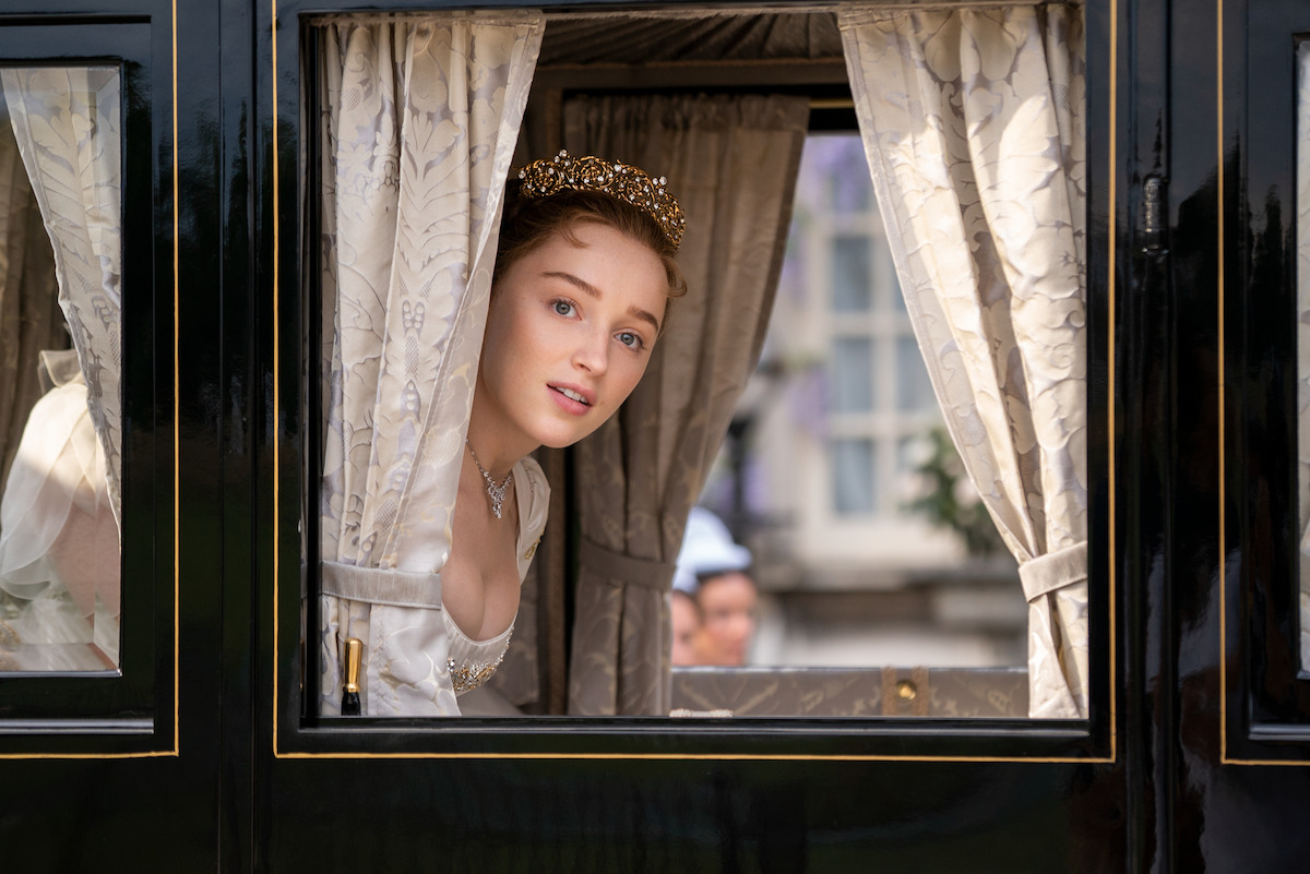 Woman in historical dress and tiara leans out of a carriage window with curtains, looking outside in a period setting with blurred figures and building in the background.