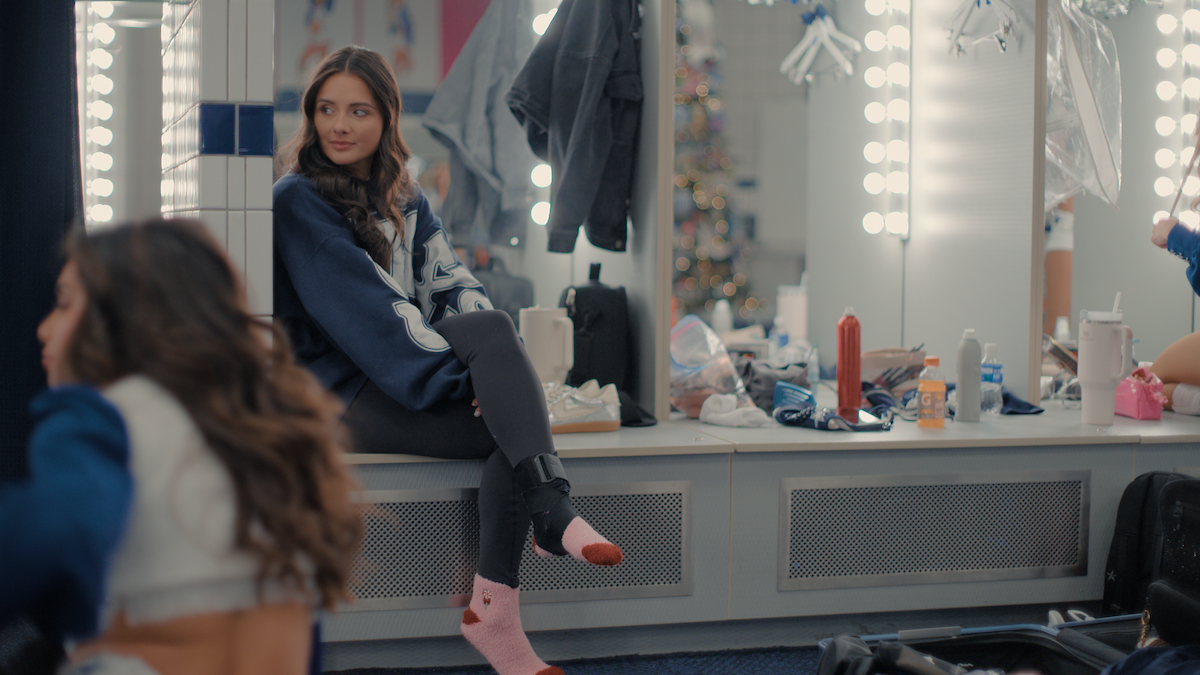 Young woman in a letterman jacket and pink socks sits on a dressing room counter, surrounded by mirrors with lights, makeup, clothes, and personal items; other people are getting ready in the lively, cluttered backstage environment.