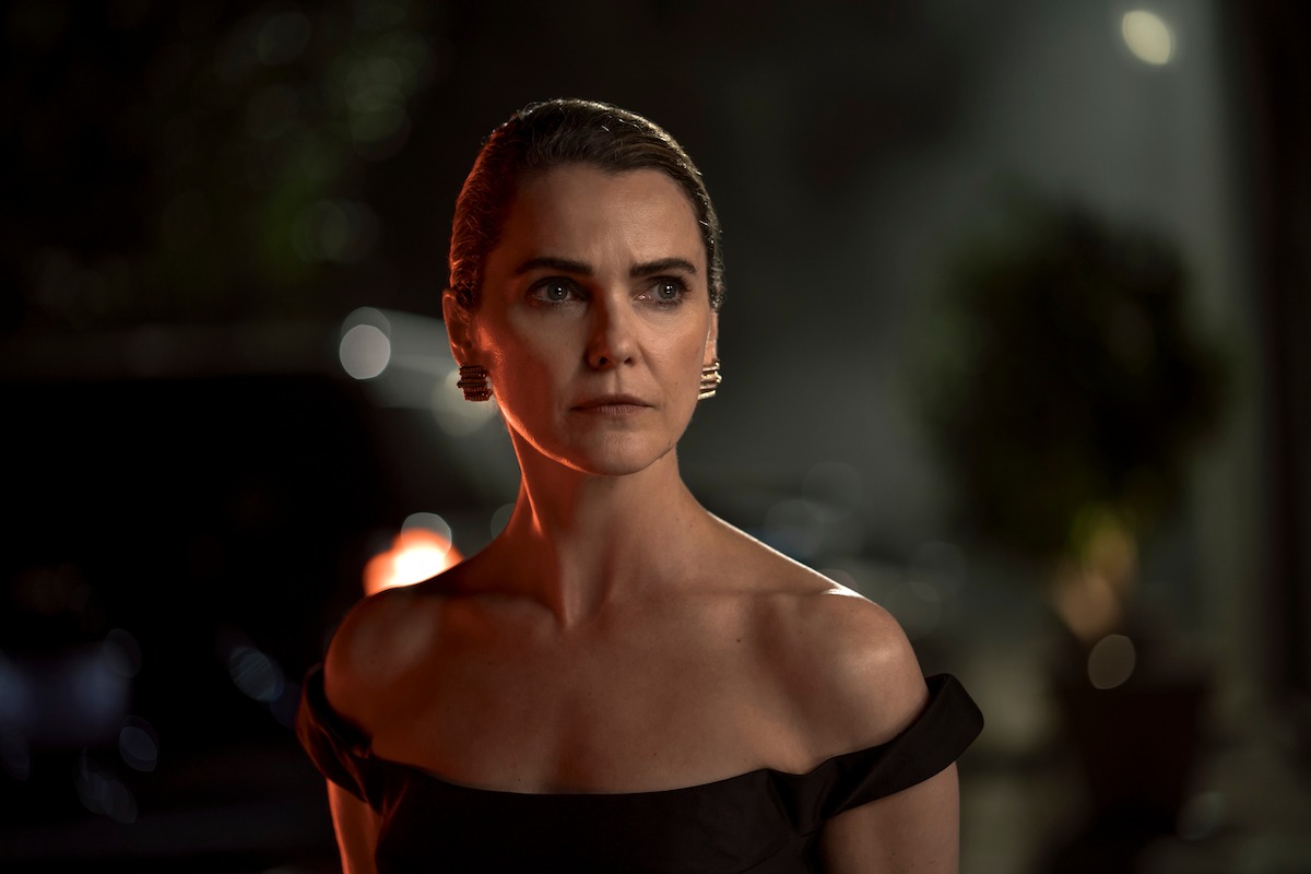 Woman with slicked-back hair and elegant black dress stands outdoors at night, dramatic lighting highlighting her serious expression, blurred background with plants and car.