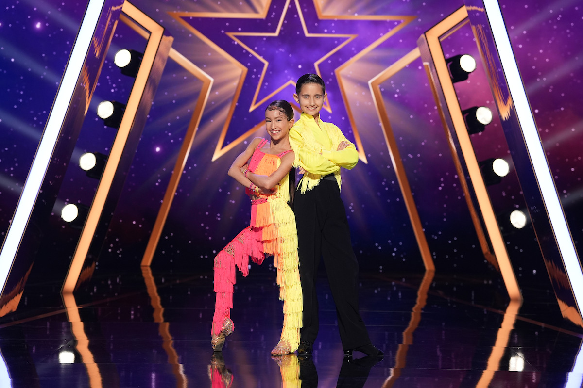 Davyd & Stella in colorful costumes pose back-to-back on a shiny stage with bright lights and a large glowing star in the background, suggesting a talent show or dance competition setting.