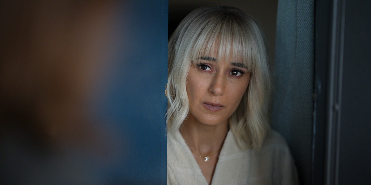 Woman with blond hair and bangs, wearing a light robe, stands in a doorway indoors, looking out with a serious expression, soft lighting and blurred blue background.