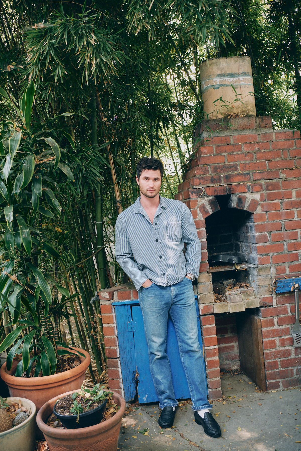 A man stands in a garden beside a brick outdoor oven, surrounded by potted plants and tall bamboo, wearing a gray shirt and blue jeans, with a relaxed atmosphere in a natural, outdoor setting.