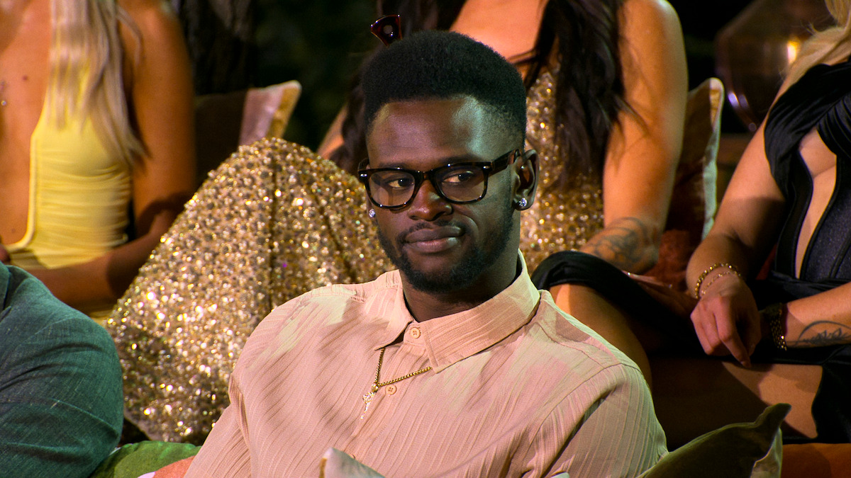 A man wearing glasses and a light shirt sits on a couch at a social event, surrounded by elegantly dressed people in eveningwear, creating a formal yet relaxed atmosphere.