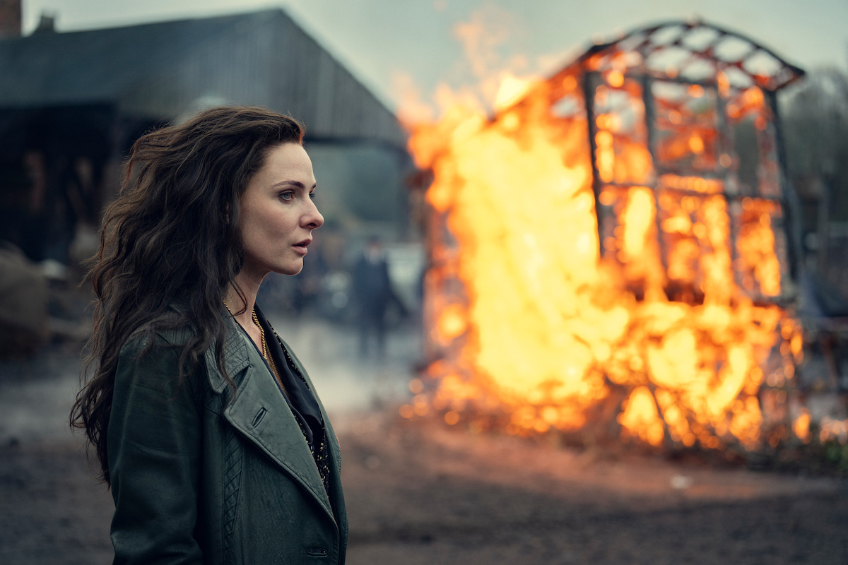 Woman in a dark coat stands outdoors with a burning wooden structure behind her; rural, overcast setting, dramatic and tense mood.