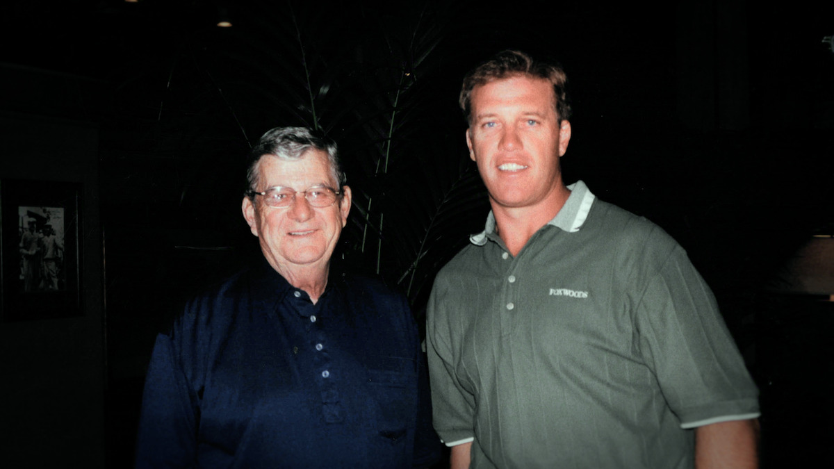 Jack and John Elway standing indoors in a dimly lit room, both looking at the camera and smiling. The man on the left wears glasses and a dark shirt, the man on the right wears a green polo. Background is dark with faint shapes visible.