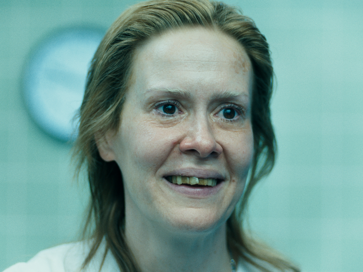 A smiling woman with light hair and visible teeth stands in a clinical setting with tiled walls and a clock in the background, under bright, diffused lighting.
