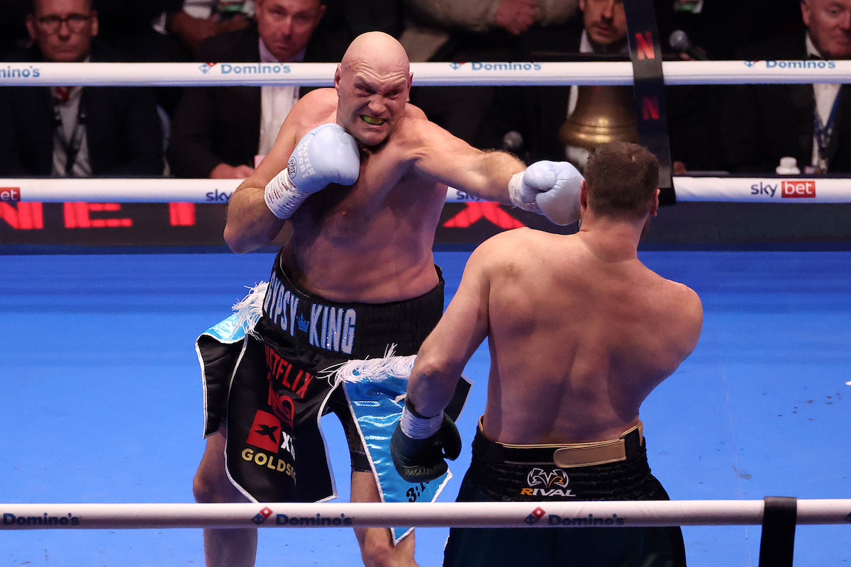 Two boxers fighting in a brightly lit boxing ring, one throwing a punch at the other, surrounded by spectators and advertising banners on the ropes.