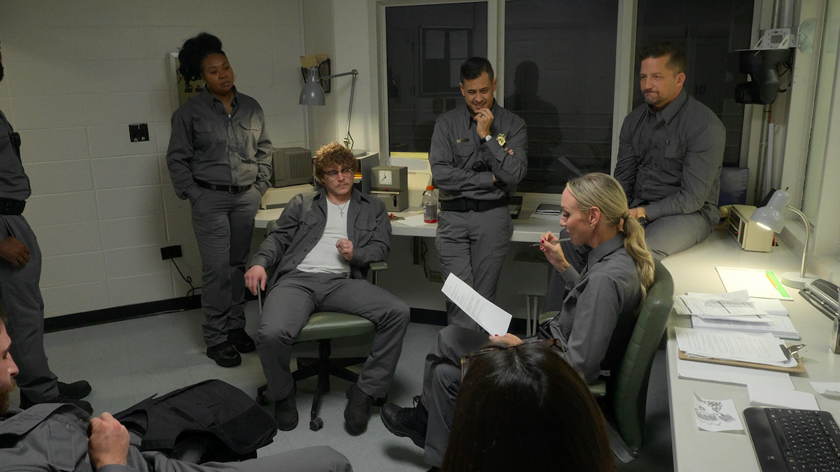 Group of seven people in gray uniforms having a discussion in a small, brightly lit office with desks, papers, and a window, suggesting a workplace or institutional setting.