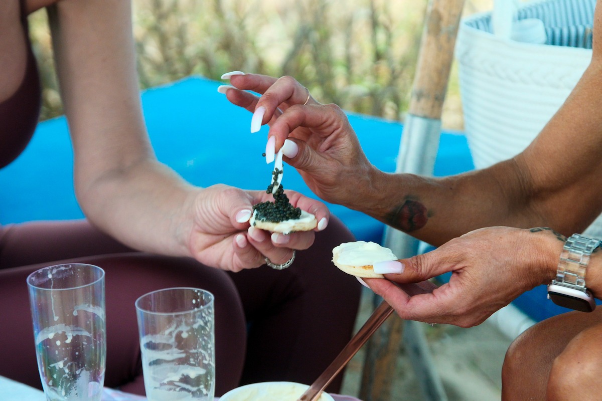 Two women at an outdoor picnic spreading caviar on crackers, with empty glasses and a relaxed, casual atmosphere. Hands and food are in focus, suggesting a moment of socializing and sharing snacks.