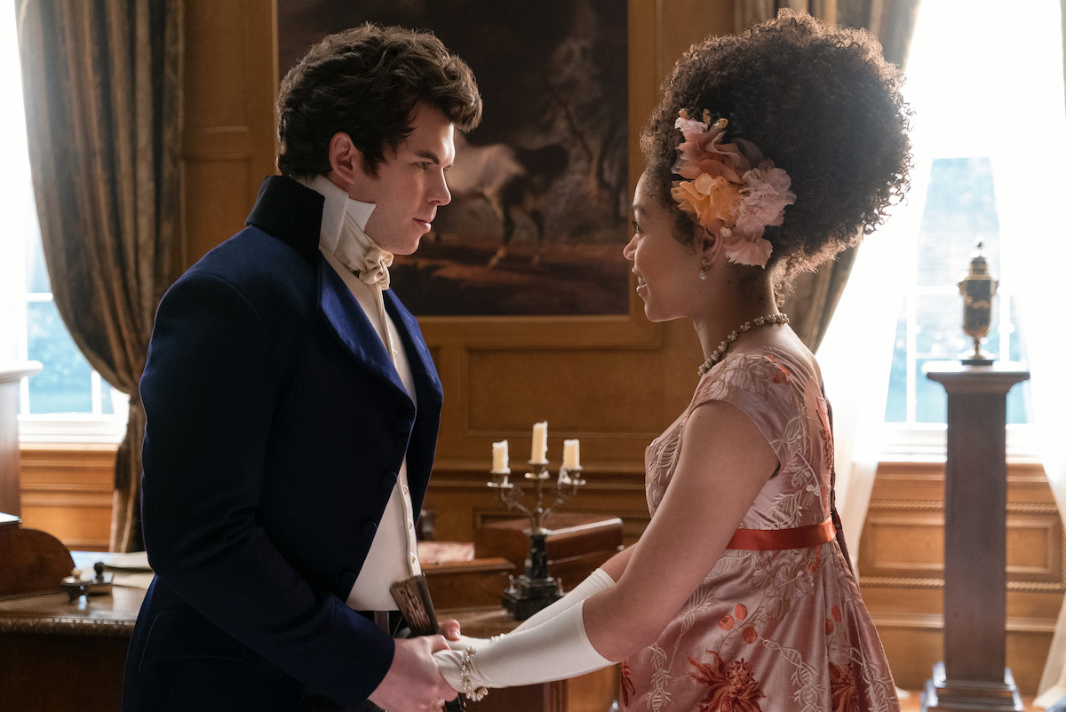 A man and woman in historical attire hold hands in an elegant, sunlit room with large windows, rich drapery, classic furniture, and a painting of a horse in the background.