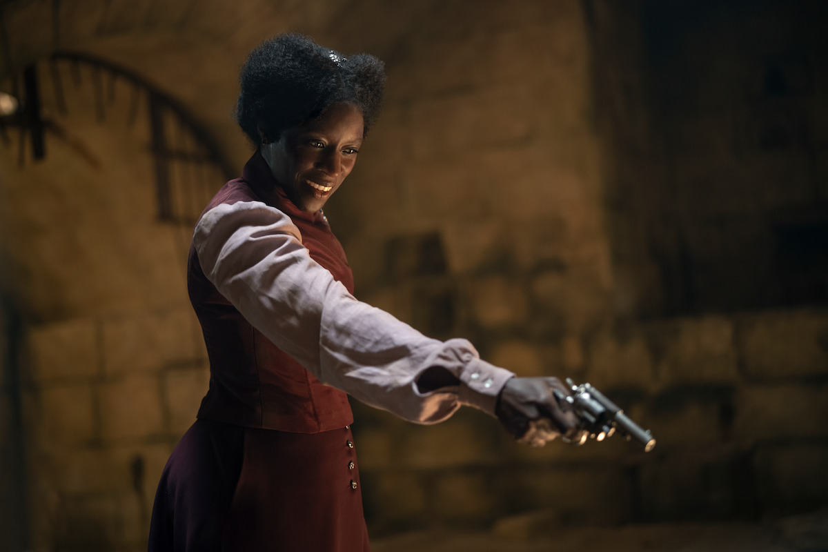 Woman in historical clothing stands in a dimly lit stone-walled room, smiling as she points a revolver, suggesting tension or drama in an old-fashioned setting.