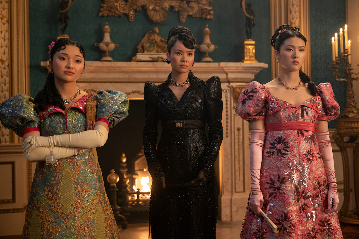Three women in elaborate, ornate gowns stand in a richly decorated room with a fireplace and candles, evoking a historical or royal setting, with elegant hairstyles and formal poses.