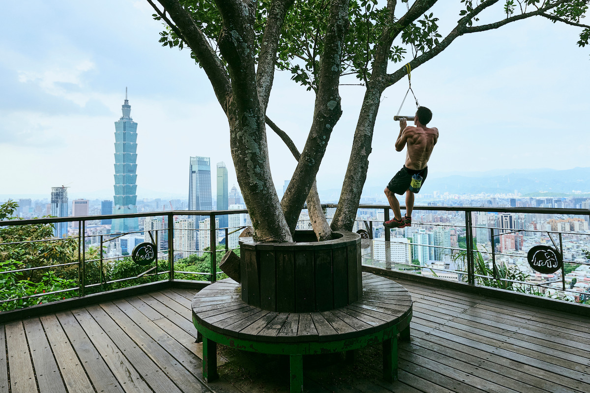 Alex Honnold swings on a rope from a tree on a wooden deck overlook with city skyline, including Taipei 101, visible in the background under a cloudy sky.