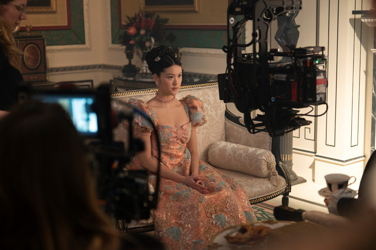 Woman in a floral ball gown sits on a vintage sofa on an ornate film set, surrounded by cameras and crew, during a period drama shoot in a richly decorated room with elegant furnishings.