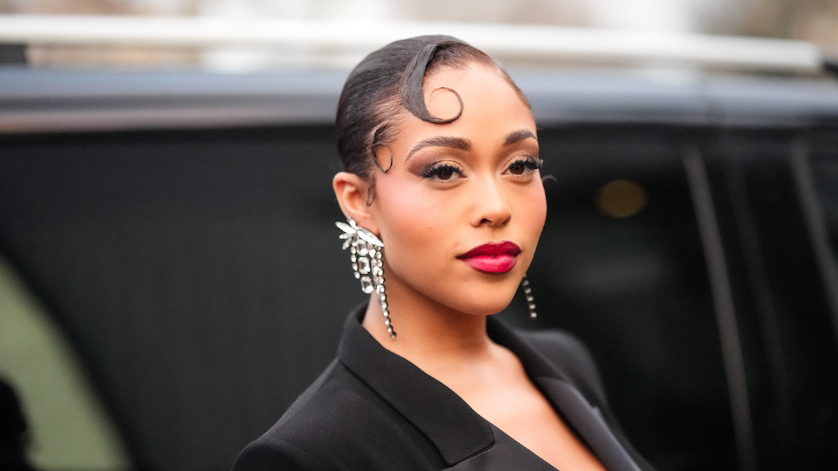 Woman with elegant hairstyle, bold makeup, and statement earrings poses outdoors wearing a black suit jacket, with a blurred black vehicle in the background.