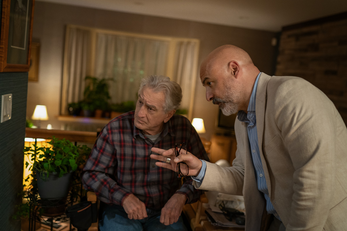 An elderly man in a plaid shirt sits indoors while another man in a beige blazer leans in, holding glasses, and speaks to him. The setting is a cozy, warmly-lit room with plants and wooden furniture in the background.