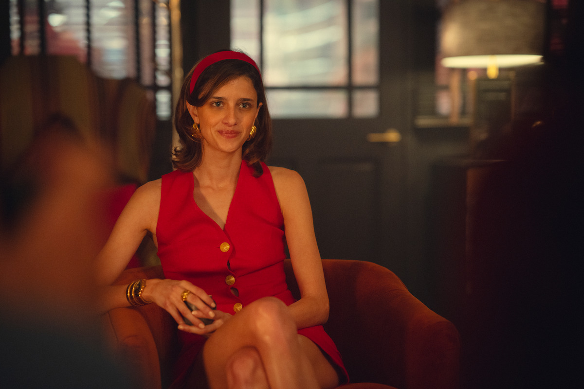 Woman in a red dress with gold buttons sits in a brown armchair, smiling in a dimly lit, cozy room with a lamp and dark doors in the background, giving a relaxed and stylish atmosphere.