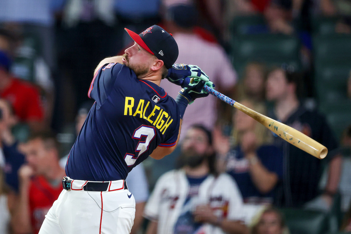 Baseball player wearing a navy jersey marked "Raleigh 3" swings a bat during a game, with a blurred crowd of fans watching in the stadium. The scene is energetic and focused on the intensity of the sport.
