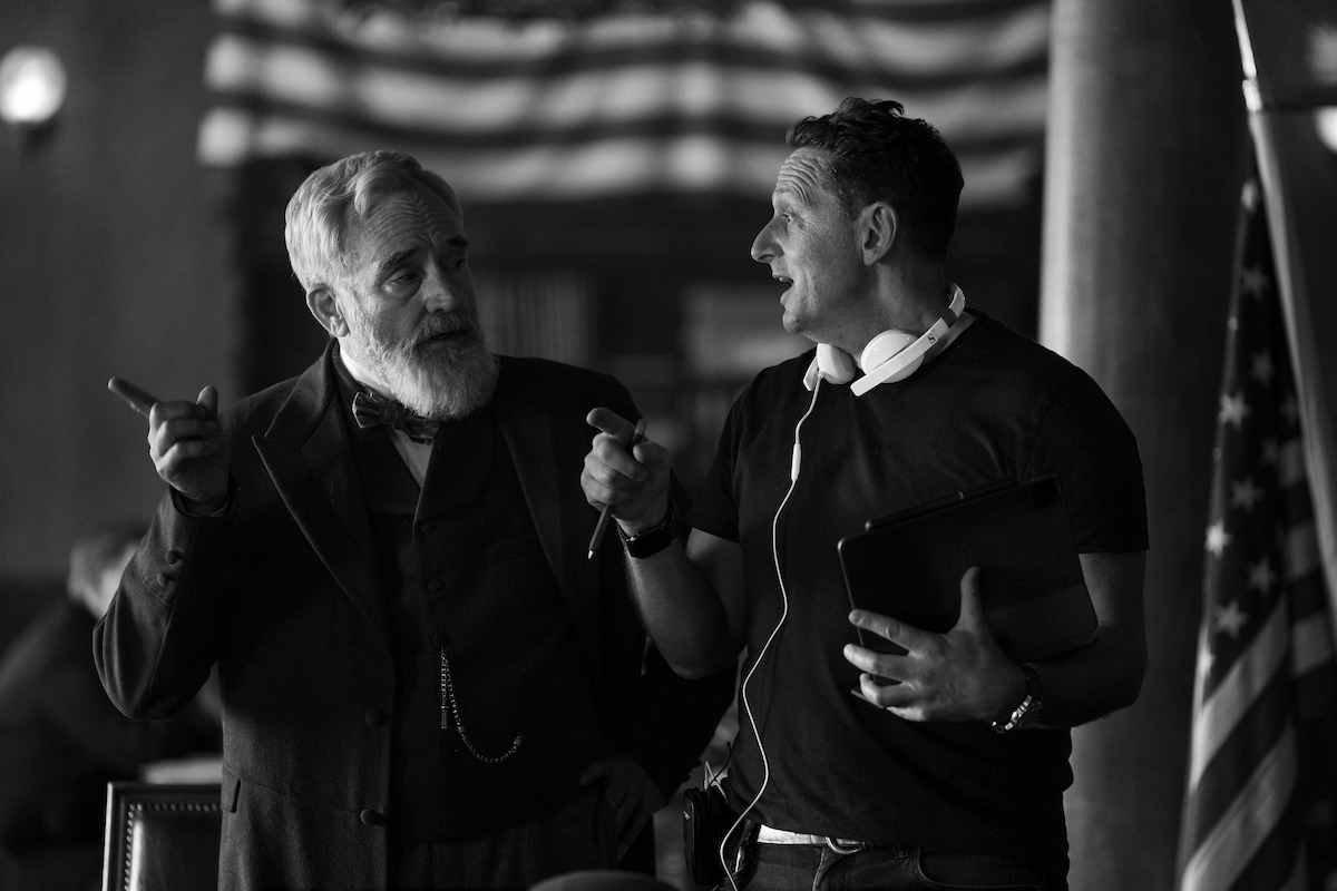 Two men in conversation indoors, one in period costume with a beard and the other in casual clothes with headphones, standing under an American flag; the mood is engaged and collaborative.
