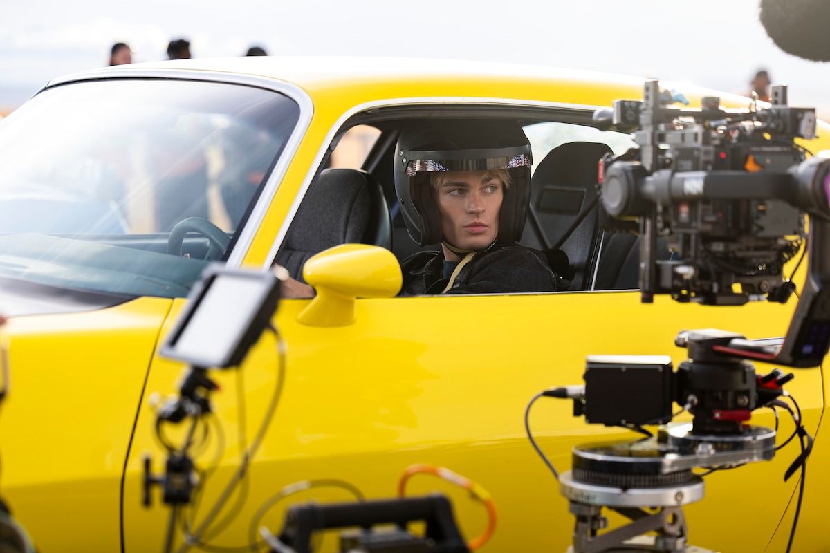 Race car driver in helmet sits in a yellow sports car during a film shoot, surrounded by professional movie cameras and equipment, creating a behind-the-scenes scene on a movie set.