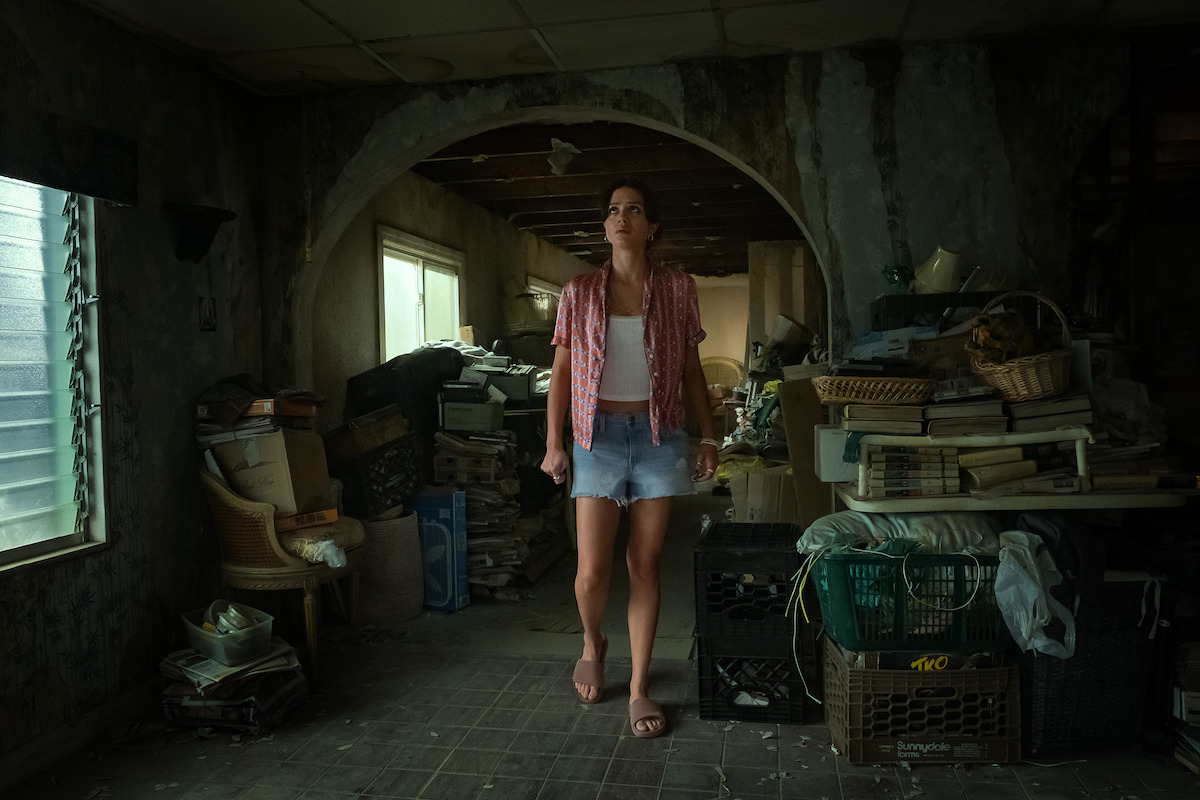 Woman in casual clothes stands barefoot in a cluttered, dimly lit room filled with old furniture, stacked books, and crates, with peeling walls and a broken ceiling, evoking a sense of neglect and disarray.