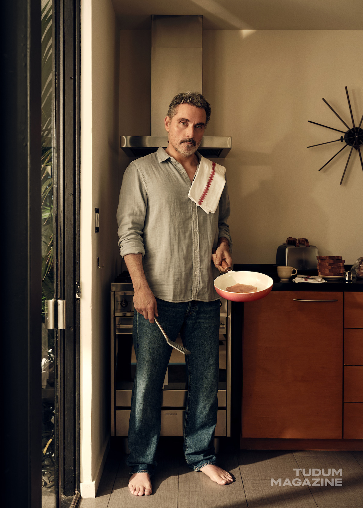 Rufus Sewell holds a frying pan in a kitchen