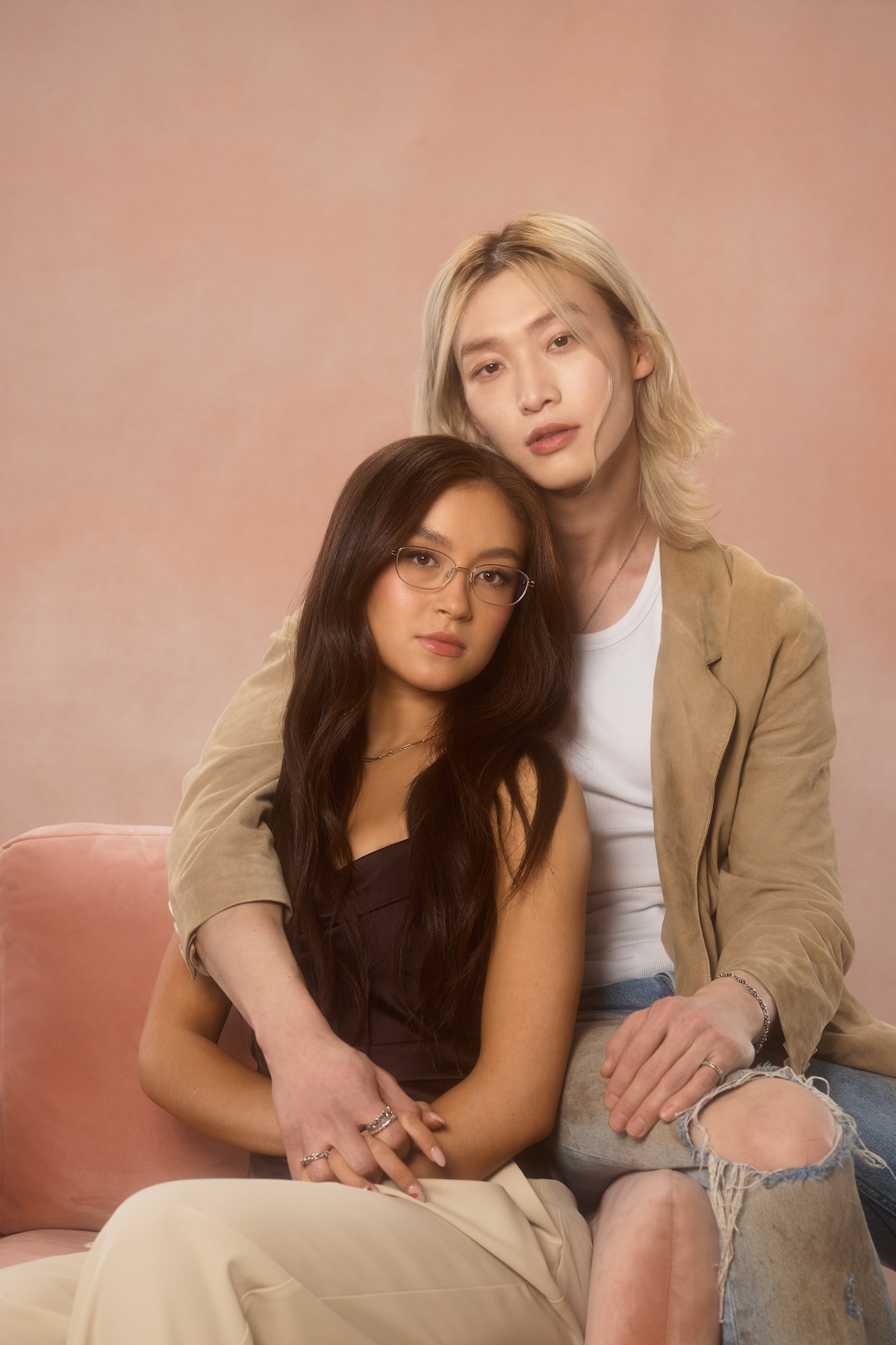 Two people sit closely on a pink sofa against a soft pink background. One wears glasses and dark top, the other has long light hair, a tan jacket, and ripped jeans. The scene feels intimate and warm, with gentle lighting.