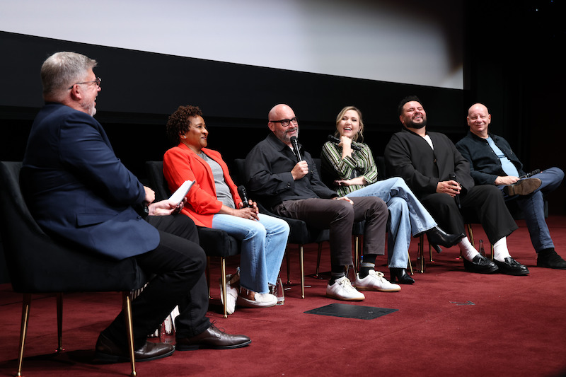 Six people seated on stage in front of a screen, participating in a panel discussion. One person holds a microphone while the others listen and smile. The setting appears to be a theater or auditorium with a red carpeted floor.