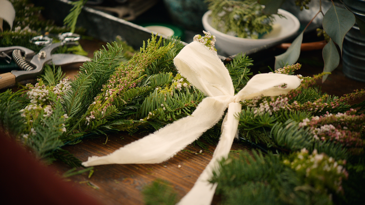 Festive evergreen wreath with small flowers and a cream ribbon bow on a rustic wooden table, surrounded by crafting tools and greenery, evoking a cozy, handmade holiday atmosphere.