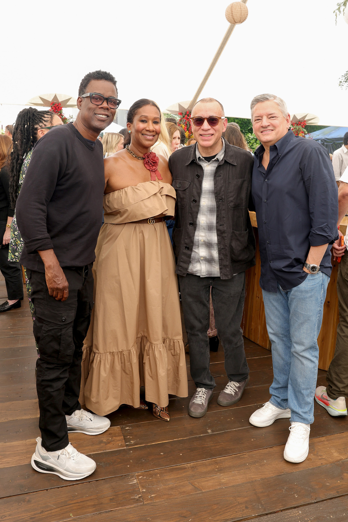Four adults pose together at an outdoor social event on a wooden deck; people and festive decorations are visible in the background, suggesting a casual, cheerful gathering.
