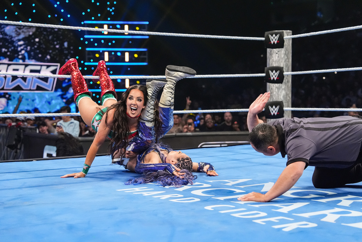 Two female wrestlers compete in a WWE ring under bright arena lights, with an excited referee counting a pin. The crowd watches the energetic match, creating a lively and intense atmosphere.