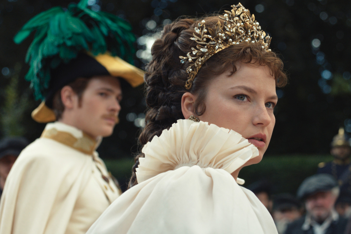 A woman in a regal white gown with a ruffled collar and a golden crown, looking serious, while a man in a feathered hat and formal attire stands blurred in the background, amidst a crowd.