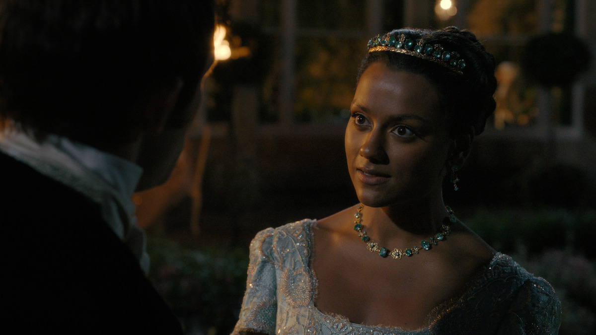 A woman in an ornate blue dress and jeweled headband stands outside at night, facing a man in formal attire. Warm lights and greenery create an elegant, historical atmosphere.