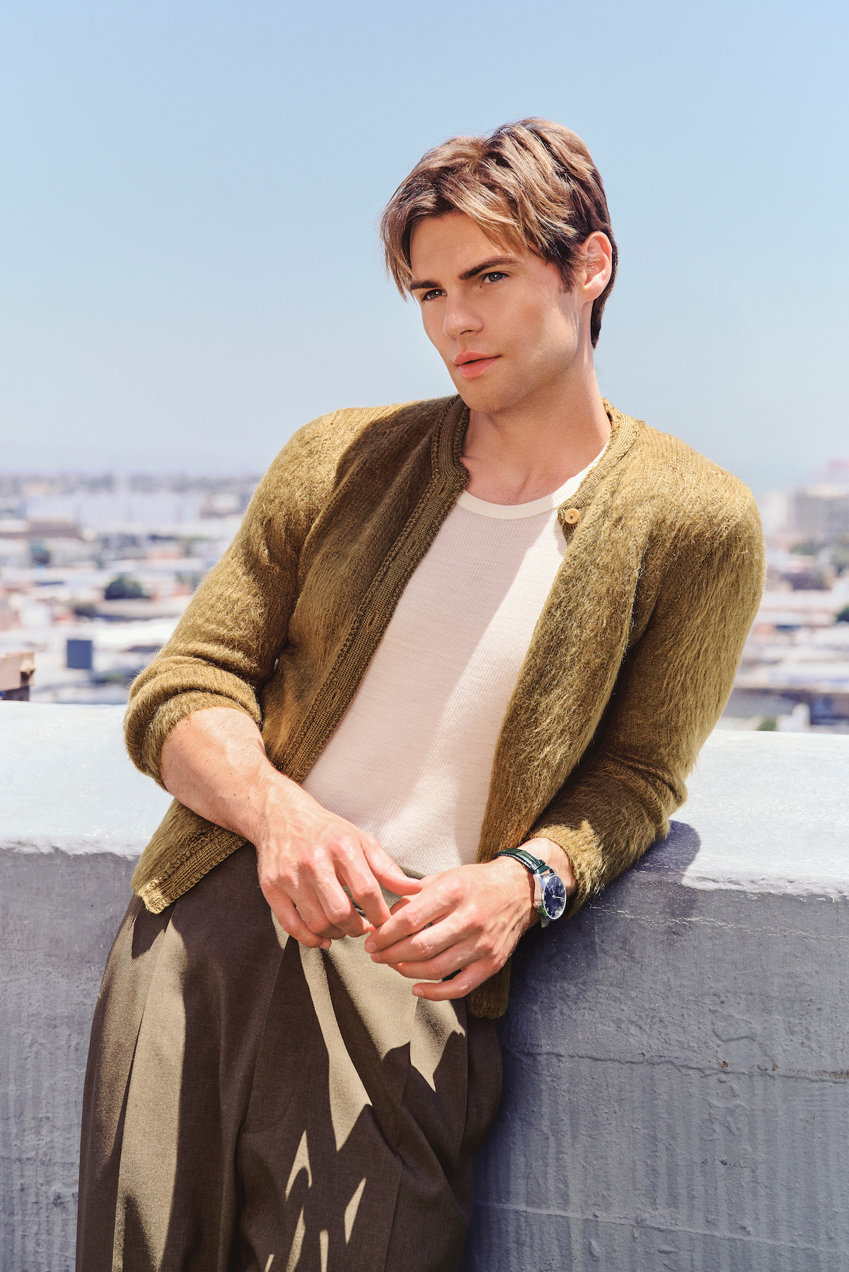 A young man in a light shirt and olive cardigan poses outdoors, leaning against a concrete ledge with a cityscape and clear blue sky in the background, suggesting a sunny urban rooftop setting.